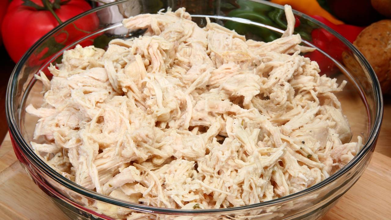 Frango desfiado, uma das opções de recheio para turbinar sua crepioca fit. Tigela de vidro com frango cozido desfiado, legumes e pão ao fundo