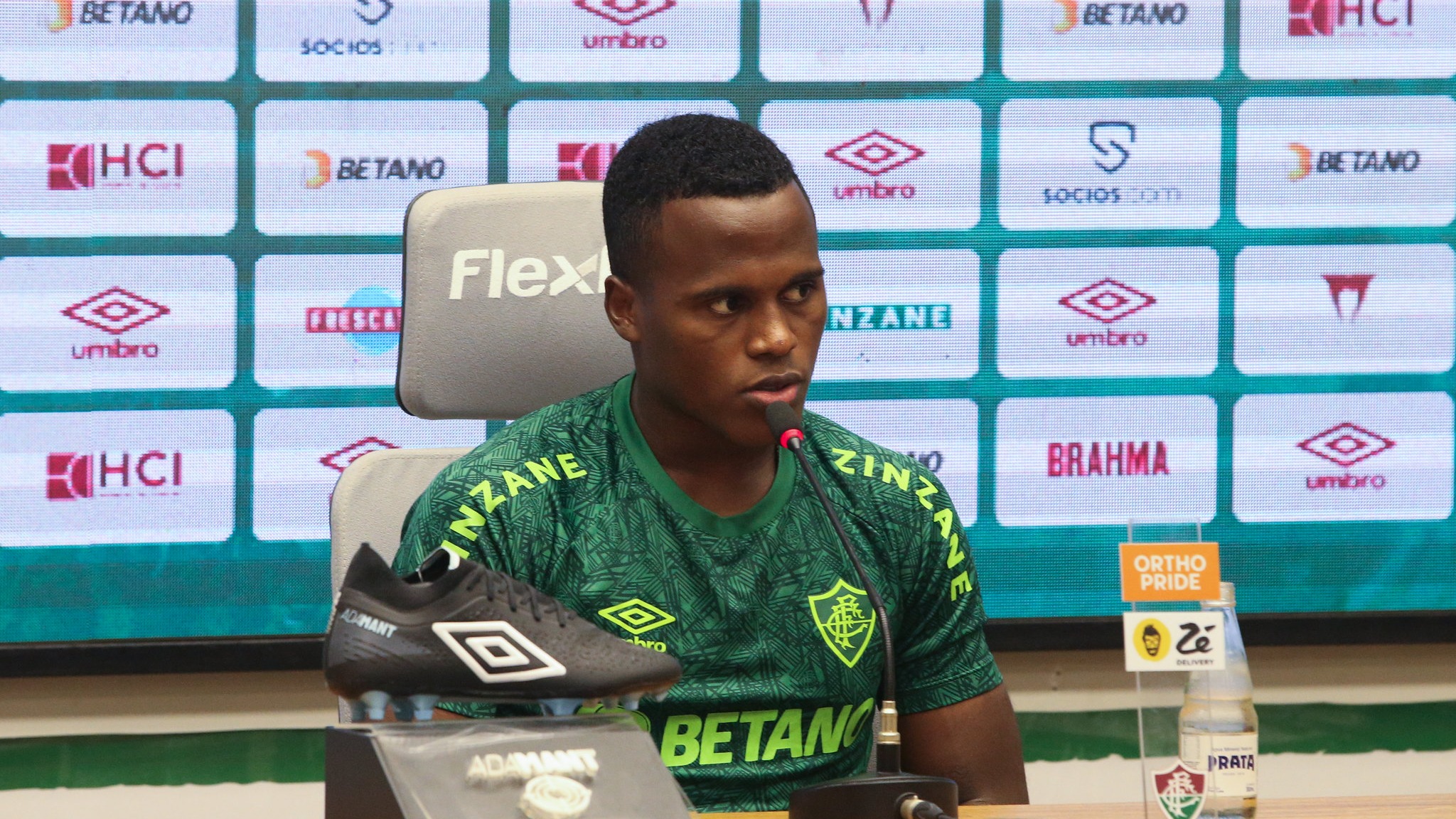 Jhon Arias em coletiva de imprensa pelo Fluminense antes de sua transferência para o Wolverhampton. Jhon Arias, camisa verde, em coletiva de imprensa com microfone e chuteira preta.