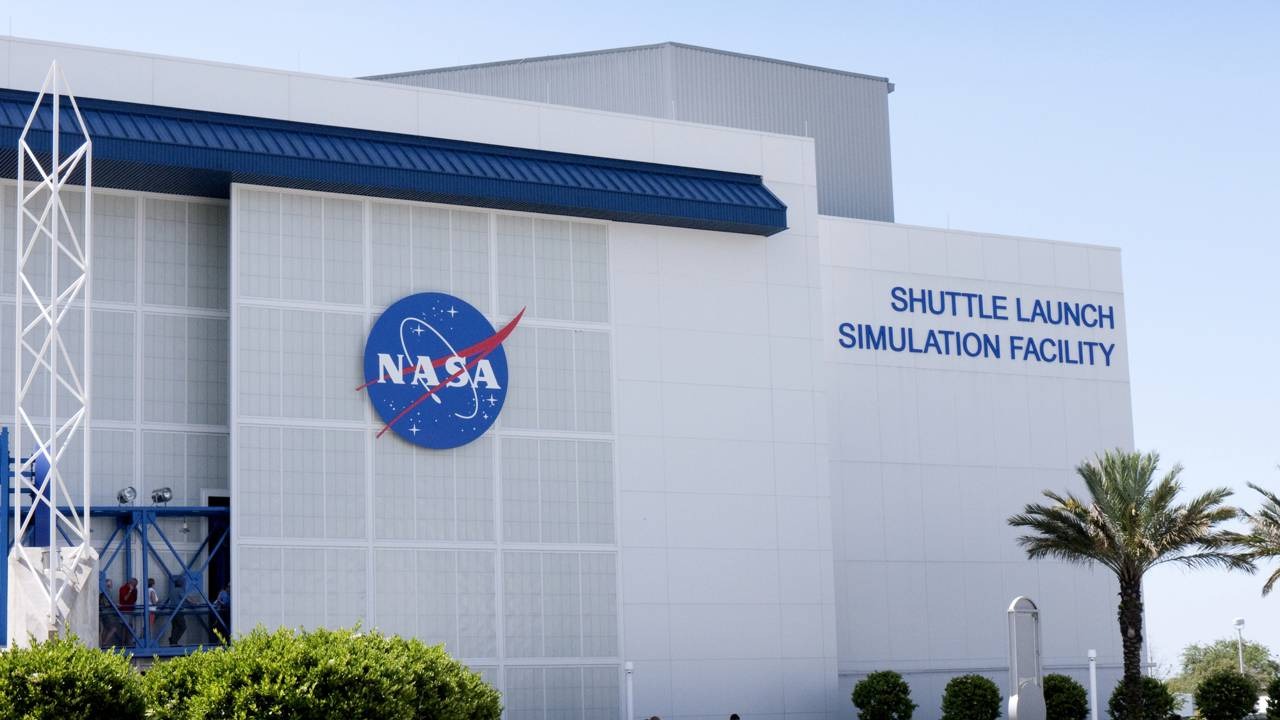 A NASA lidera pesquisas sobre asteroides como Bennu, fundamentais para entender a origem da vida. Edifício da NASA com logo, "SHUTTLE LAUNCH SIMULATION FACILITY", palmeiras e arbustos