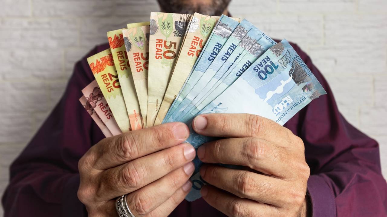 O debate sobre o reajuste do salário mínimo e seus efeitos na economia brasileira impacta diretamente o valor no bolso dos trabalhadores. Créditos: depositphotos.com / verganifotografia Mãos seguram notas de Real brasileiro: 20, 50 e 100 Reais.