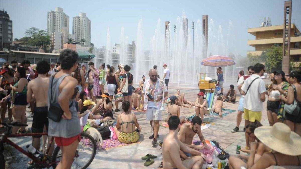 Multidão em roupas de verão se reúne em praça urbana com fontes d'água e prédios ao fundo.