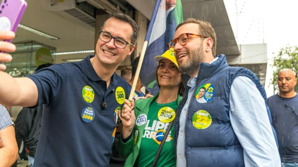 Deltan Dallagnol e Filipe Barros, senadores eleitos pelo Paraná, em registro de campanha antes da confirmação de suas vitórias. Foto: Divulgação Homens e mulher sorriem em selfie, usando broches de campanha 'Deltan' e 'Filipe'