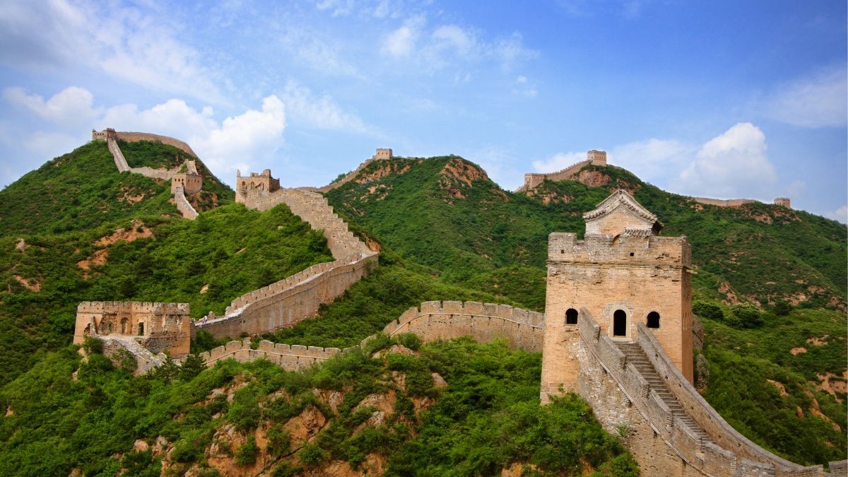 A Grande Muralha da China, um símbolo da persistência e grandiosidade da civilização, figura entre as Sete Maravilhas do Mundo Moderno. A Grande Muralha da China serpenteando por montanhas verdes sob céu azul com nuvens.