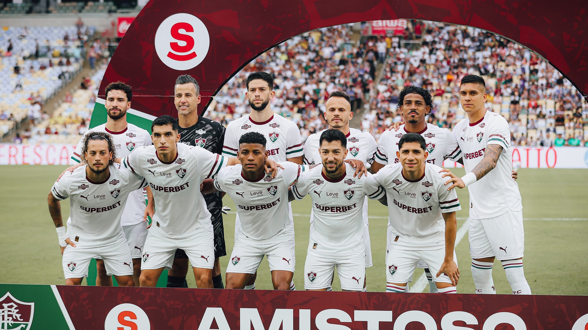 O elenco do Fluminense se apresenta para o retorno do Campeonato Brasileiro, com a expectativa dos novos reforços e a base da equipe FOTO: LUCAS MERÇON / FLUMINENSE F.C. Onze jogadores do Fluminense posam uniformizados em campo, com patrocinadores visíveis e arco S