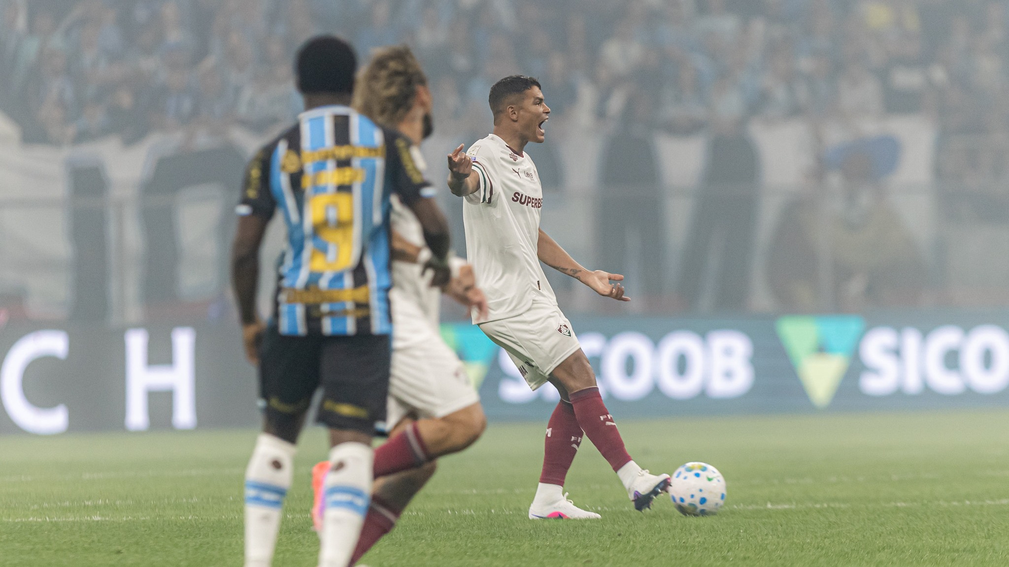Jogador do Fluminense demonstra intensidade em campo, refletindo a atmosfera dos jogos decisivos que o clube enfrentará nas próximas semanas Jogador de camisa branca e bordô grita no campo com bola, dois jogadores embaçados e torcida ao fundo