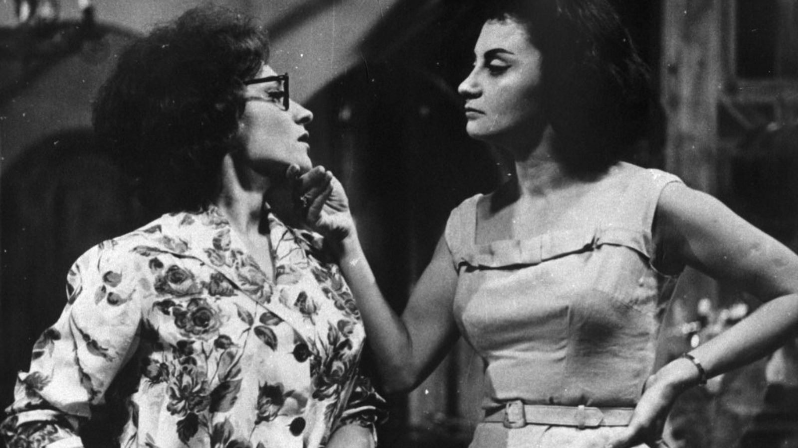Duas jovens atrizes em cena dram&aacute;tica, uma com &oacute;culos e blusa floral, outra com vestido liso