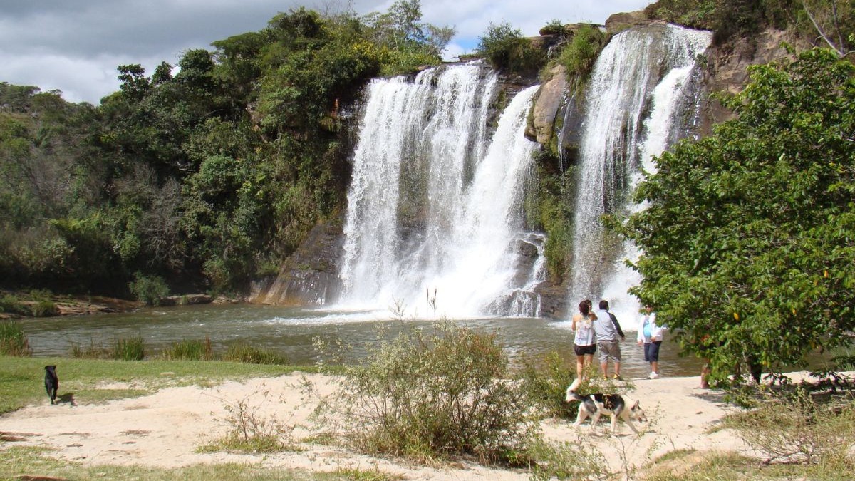 Cachoeira em Minas Gerais com pessoas e cachorros na margem do rio, rodeada por natureza.