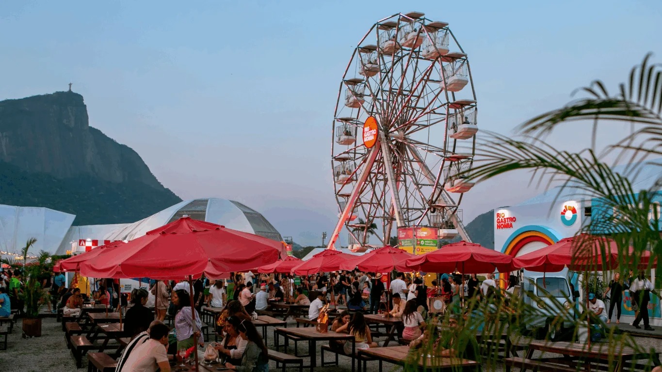 Último fim de semana do Rio Gastronomia no Jockey Club Brasileiro atrai público para a festa culinária e de lazer. Foto: Divulgação Evento Rio Gastronomia: pessoas, roda-gigante, mesas, guarda-sóis e Cristo Redentor ao fundo