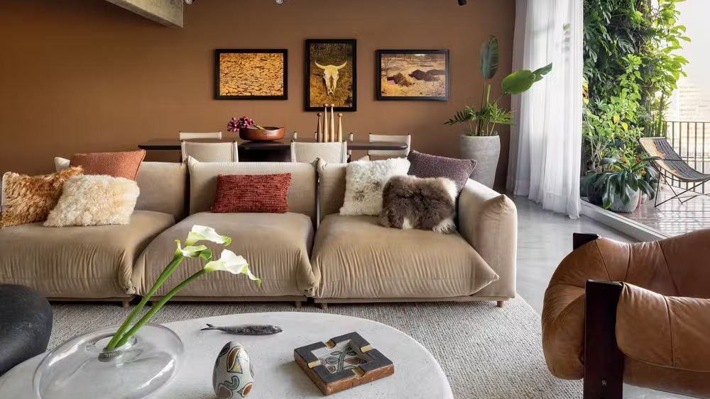 Sala de estar: sofá bege, almofadas texturizadas, mesa de centro, plantas e quadros em parede marrom