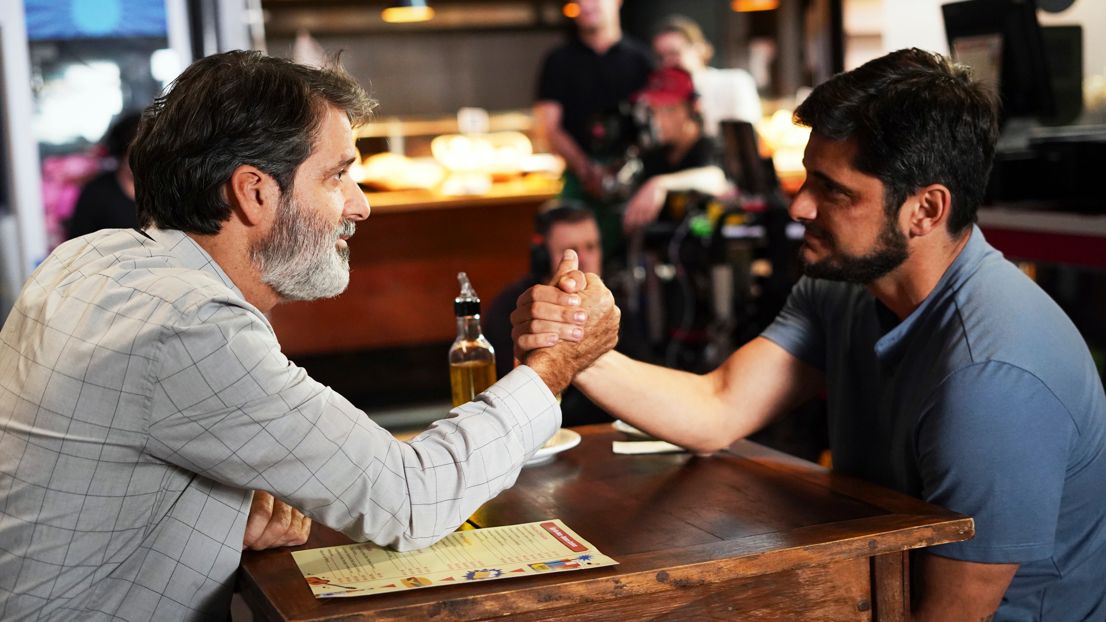 Dois homens fazem queda de braço em mesa de madeira, focados na disputa, em restaurante.