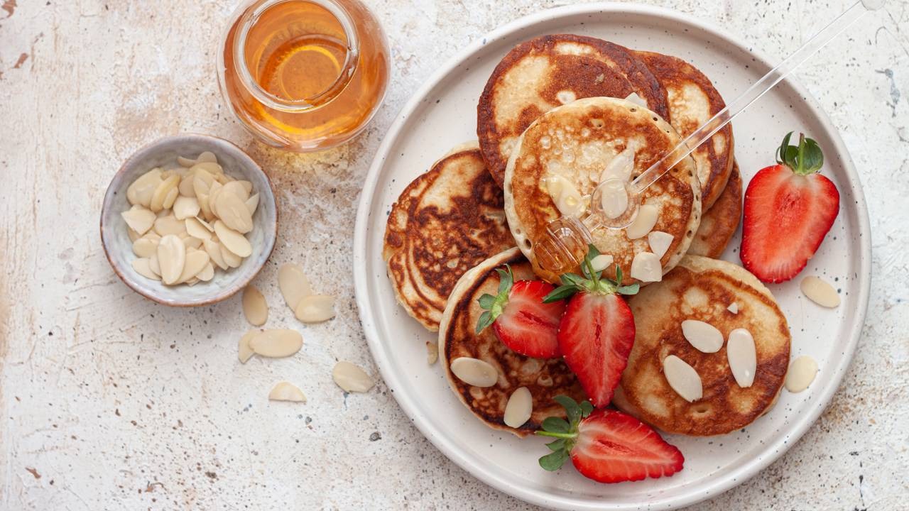 Panquecas de farinha de amêndoas com frutas e mel são a sugestão sem glúten para um café da manhã nutritivo de terça-feira. Mini panquecas com morangos, amêndoas, mel, jarra de mel e tigela de amêndoas