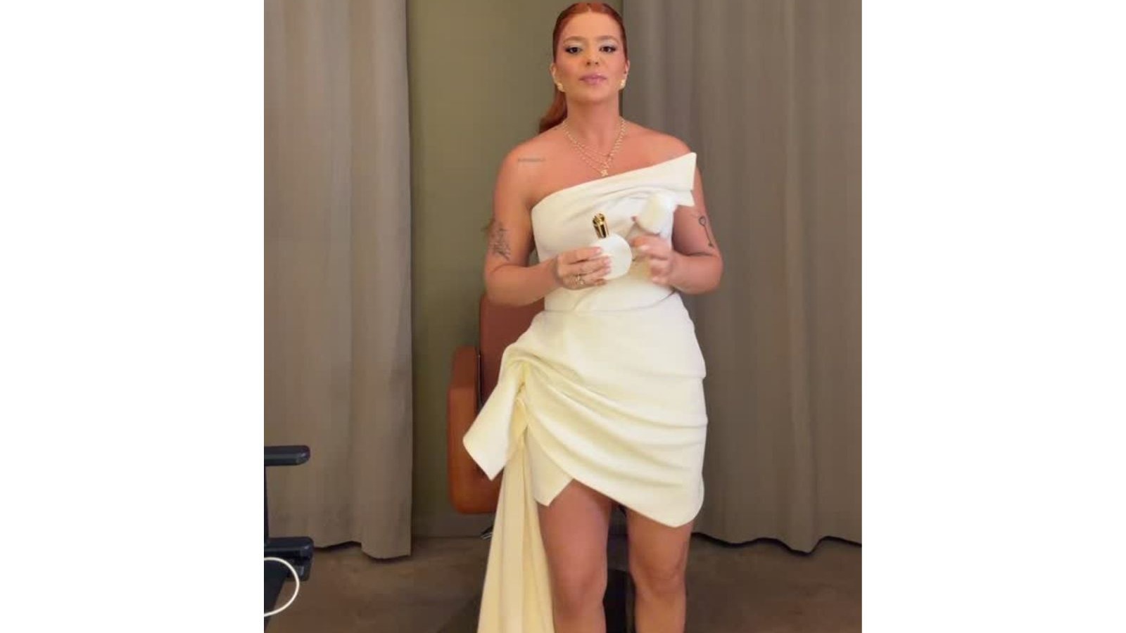 Mulher ruiva em vestido branco curto e drapeado, segurando frasco de perfume branco