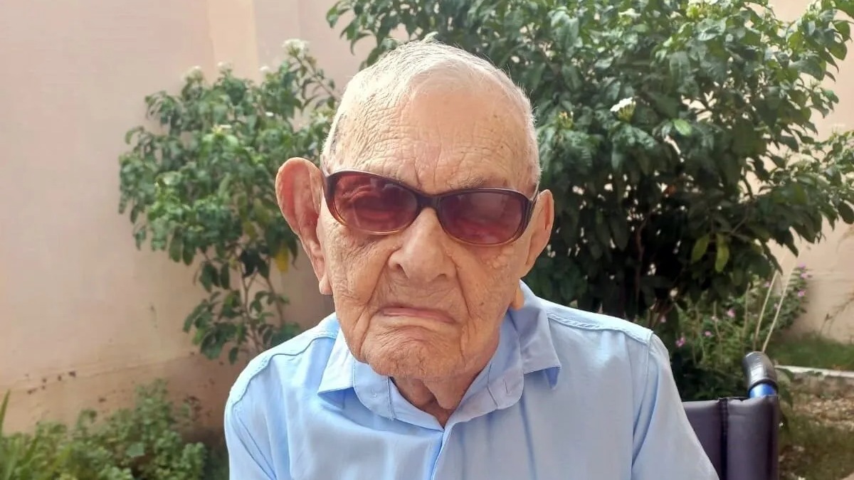 João Marinho Neto, o homem mais velho do Brasil e entre os mais longevos do mundo, celebra seus 114 anos. João Marinho Neto, homem idoso, óculos escuros, camisa azul, sentado em cadeira de rodas com plantas.