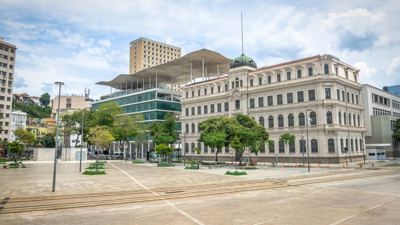 A Praça Mauá, um dos cartões-postais do Rio, oferece cultura e vistas espetaculares sem custo, um dos roteiros econômicos da cidade. Praça urbana no Rio de Janeiro com prédios históricos e modernos, árvores e trilhos no chão.