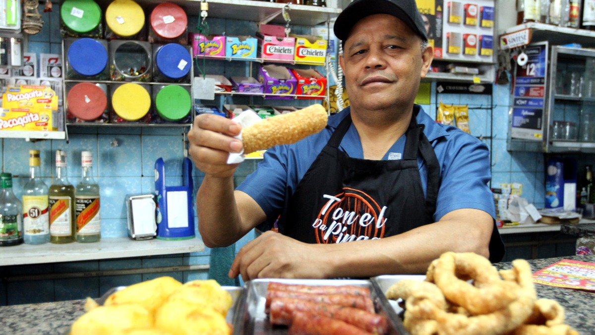Homem com avental Tonel da Pinga exibe salgado frito em balc&atilde;o de bar.