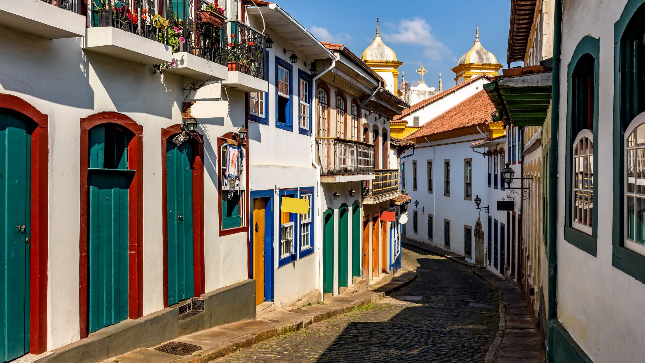 As ruas de pedra dos centros históricos mineiros, como Ouro Preto, oferecem um cenário ideal para romances à moda Bridgerton. Rua de pedra ladeada por casarões coloniais coloridos e cúpulas de igrejas sob sol.