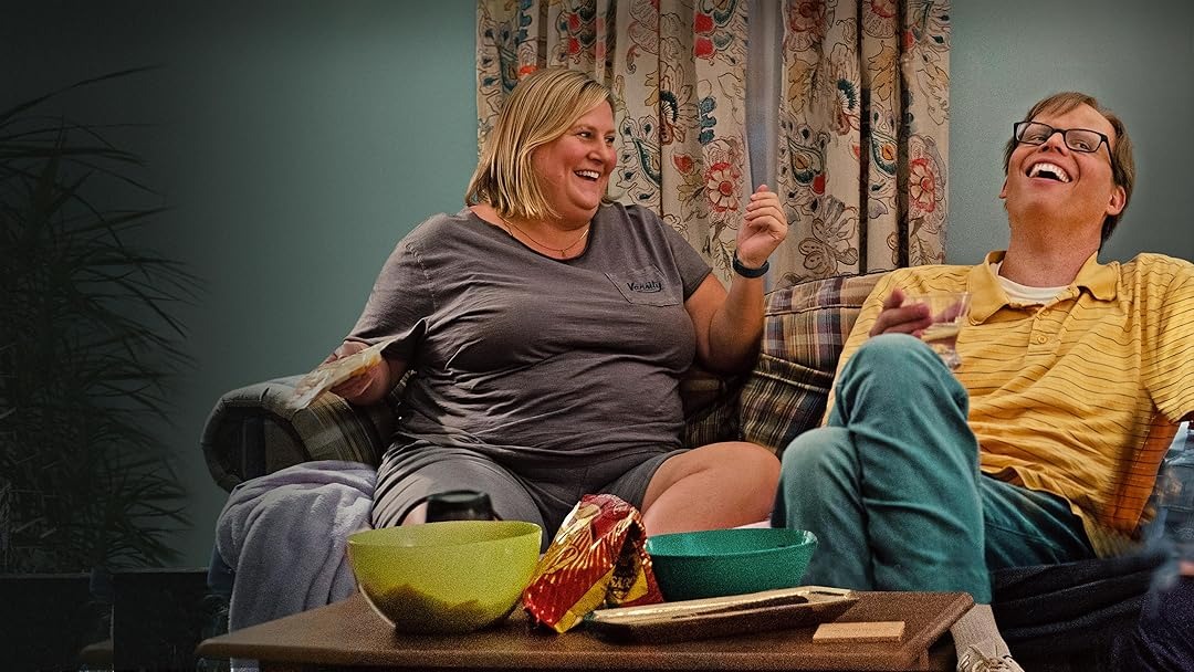 Mulher e homem rindo no sofá com tigelas e lanches na mesa de centro.