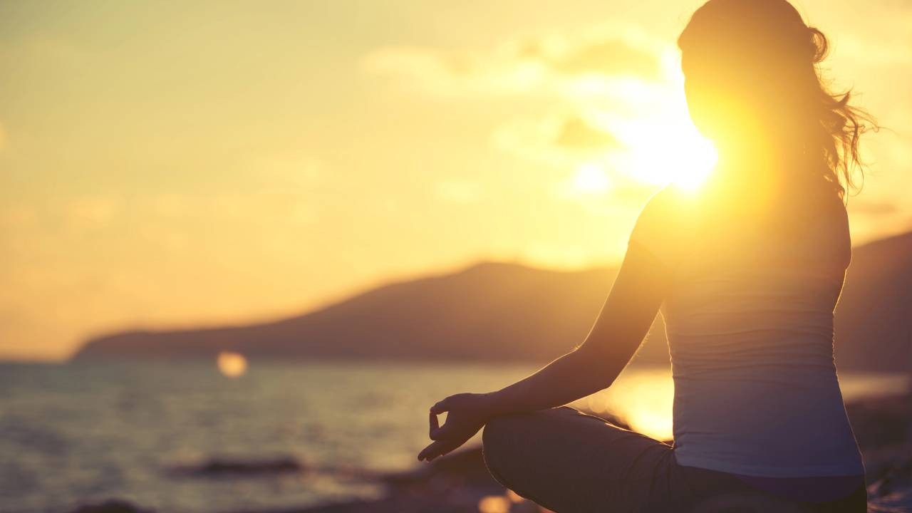 A prática de meditação ao amanhecer, um dos pilares da rotina matinal de Gisele Bündchen para nutrir mente e corpo. Silhueta de pessoa meditando em posição de lótus ao amanhecer na praia com montanhas e mar