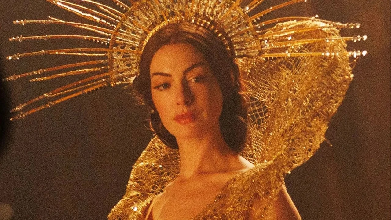 Anne Hathaway no filme Mother Mary. Anne Hathaway em close-up, com elaborada coroa dourada em formato de halo e vestes cintilantes.