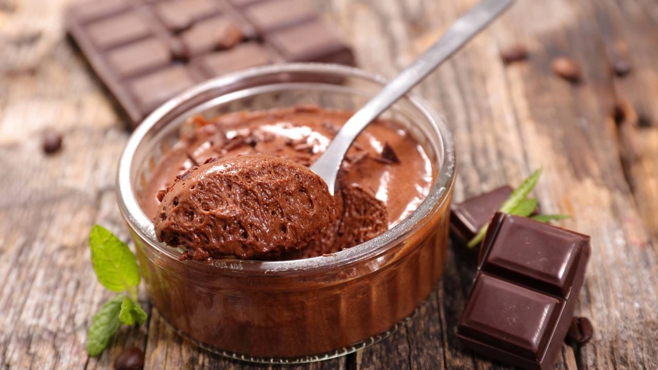 A mousse de chocolate, fácil e rápida de fazer com apenas quatro ingredientes. Tigela de mousse de chocolate com colher, barras de chocolate, grãos de café e folhas de hortelã.