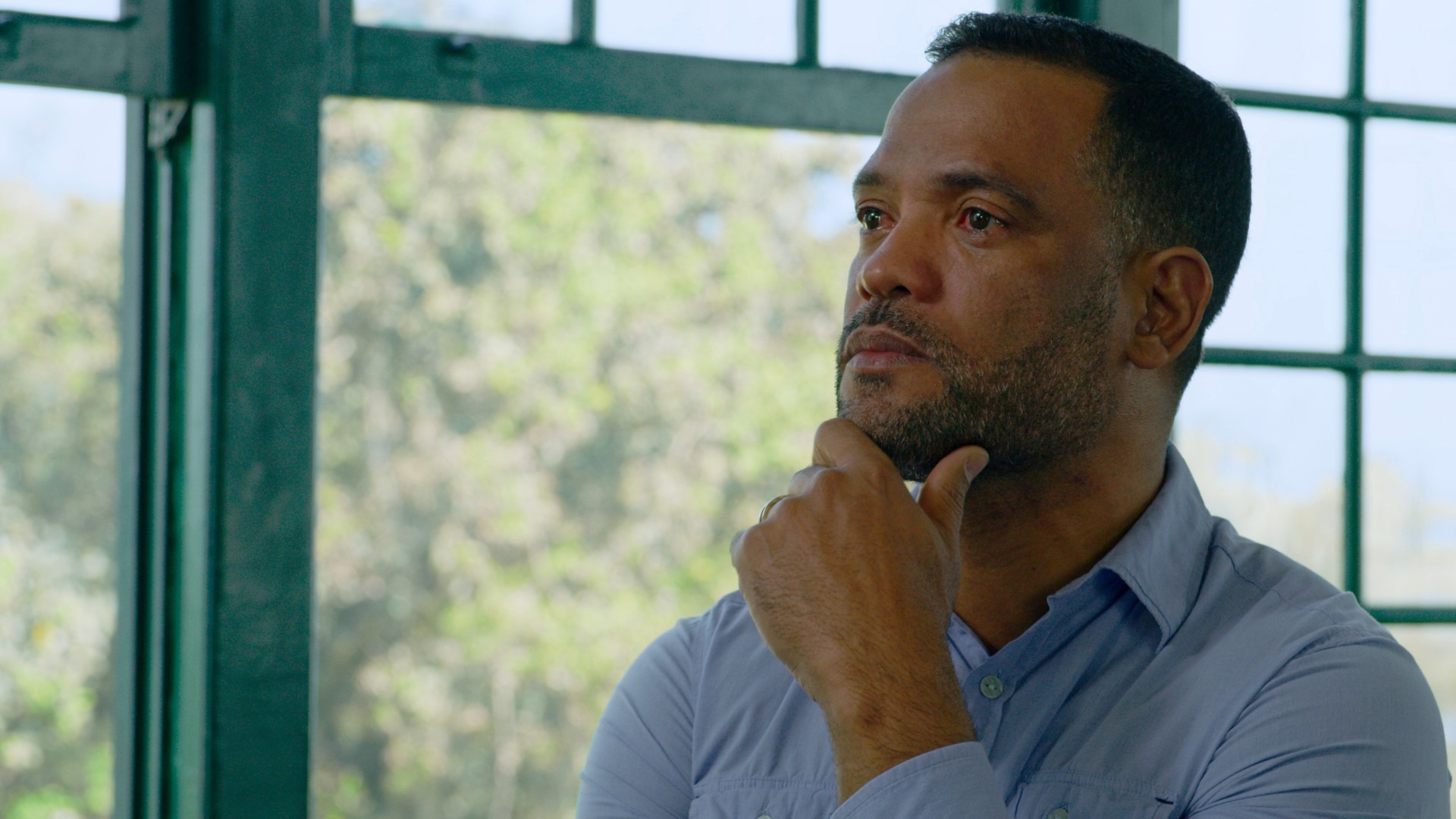 A complexidade das histórias de true crime no Prime Video convida à profunda reflexão sobre crimes reais e suas ramificações. PCC: Poder Secreto. Homem barbudo em camisa azul, pensativo, mão no queixo, olhando pela janela com grades.
