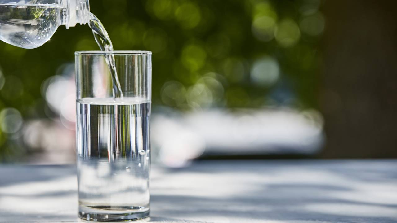 A qualidade da água que chega às residências pode ser alterada após manutenções; saiba como agir para garantir sua segurança. Água cristalina sendo despejada de garrafa plástica em copo de vidro sobre mesa clara.