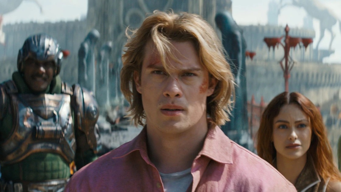 Nicholas Galitzine (centro) como Príncipe Adam, o He-Man, em cena do aguardado filme live-action de "Mestres do Universo". Nicholas Galitzine como He-Man com cabelo loiro, ladeado por homem em armadura e mulher.