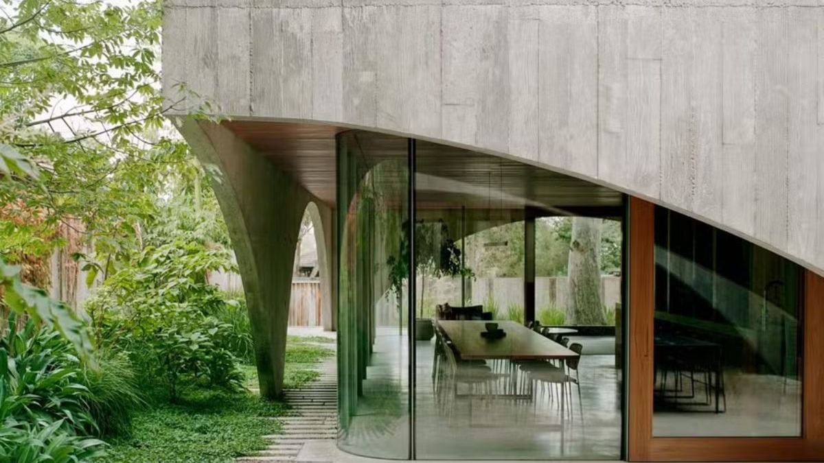 A Hawthorn House, cenário de "All Her Fault" Fachada de casa moderna com concreto curvo, paredes de vidro, jardim verde e mesa de jantar interna.