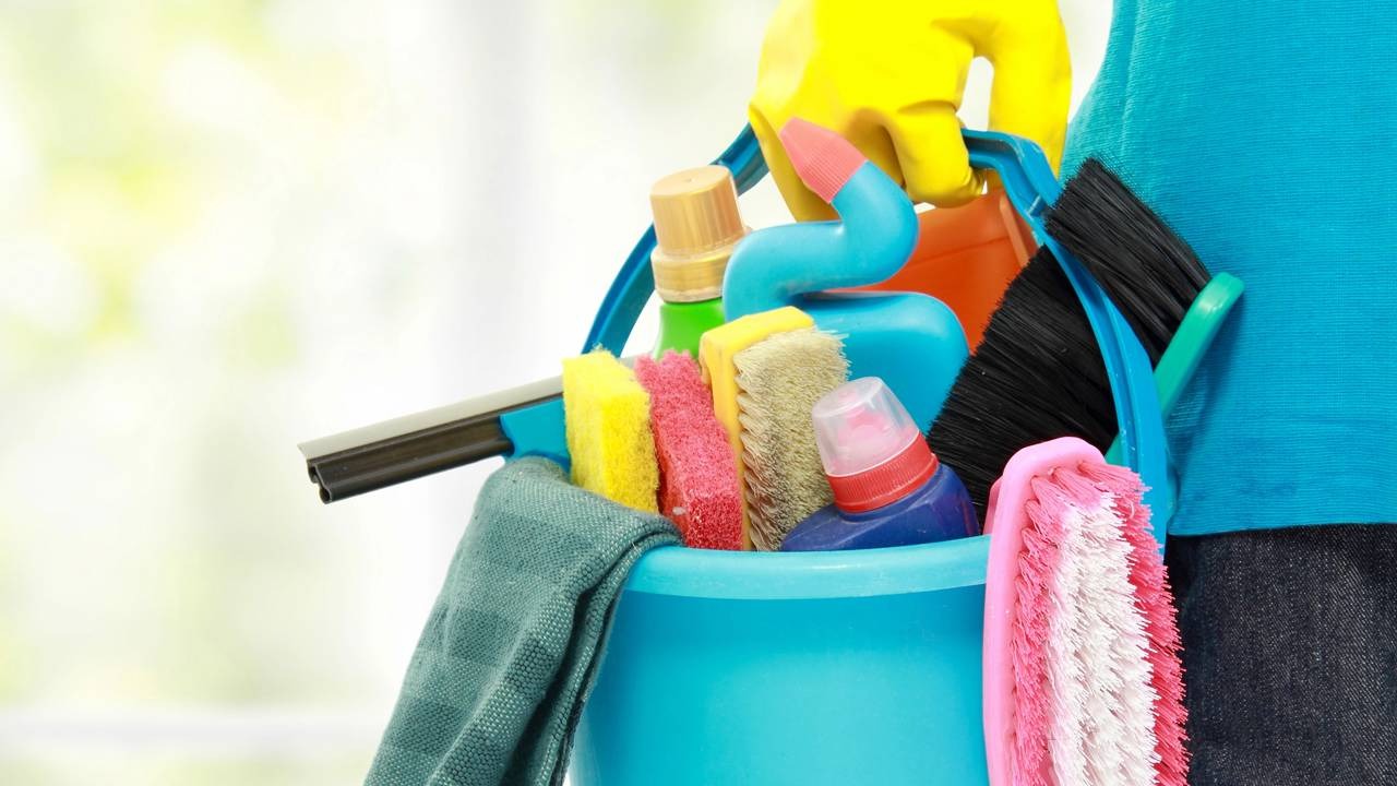 A organização prévia da casa, com os produtos certos, otimiza o trabalho da limpeza profissional e o tempo de todos. Mão enluvada amarela segura balde azul com esponjas, escovas, rodo, produtos de limpeza e pano