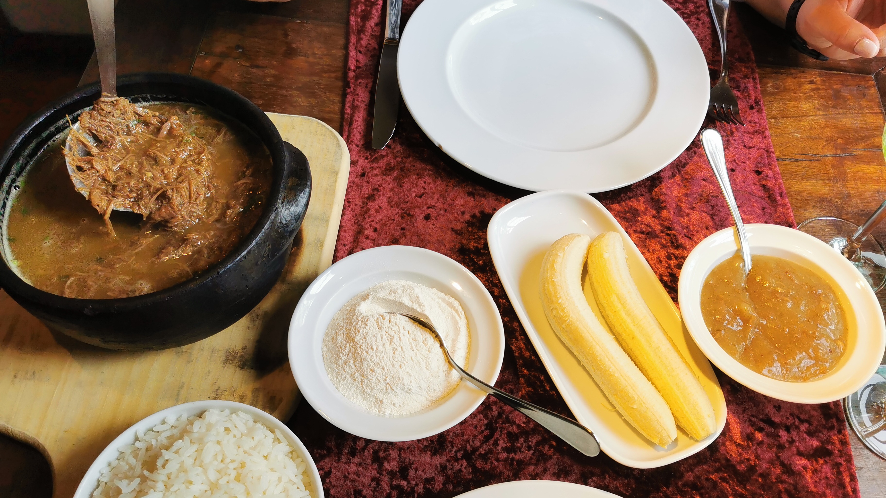 O tradicional barreado, prato típico do litoral paranaense, é cozido por horas em panela de barro e servido com farinha de mandioca e banana. Panela com barreado de carne desfiada, arroz, farinha, bananas descascadas e molho na mesa.
