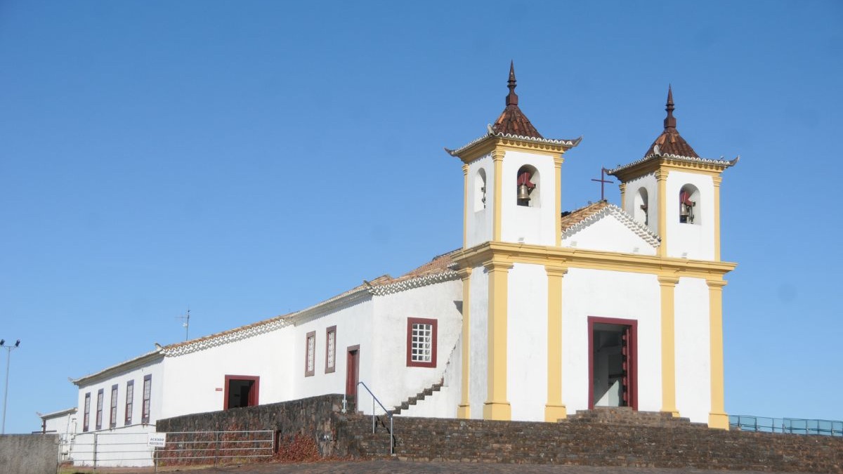 Igreja colonial branca com torres sineiras, detalhes amarelos e escadaria de pedra, c&eacute;u azul.