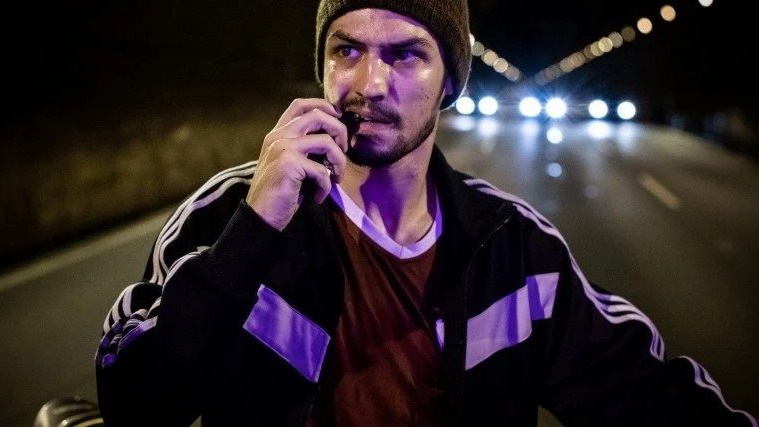 Série brasileira 'Dom' conta uma história real. Homem sério com gorro e jaqueta esportiva, falando em rádio, em ambiente noturno