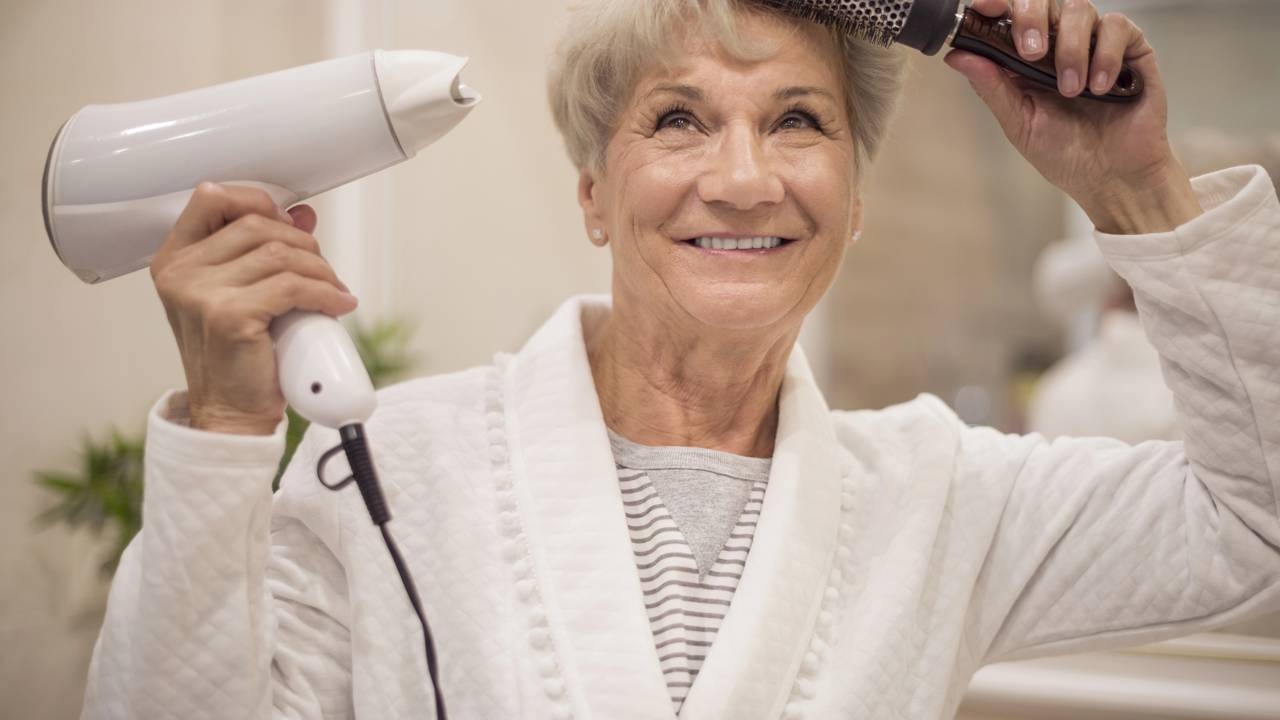 Técnicas adequadas de secagem e estilização complementam os cuidados para cabelos grisalhos e coloridos. Mulher idosa sorridente seca o cabelo grisalho com secador e escova