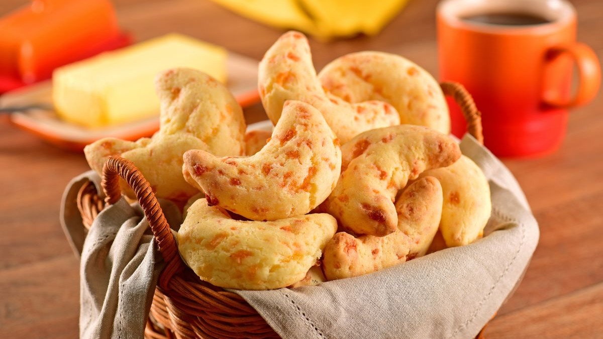 A chipa representa a forte influência da culinária paraguaia nos sabores autênticos de Mato Grosso do Sul. Cesta de chipas em formato de ferradura, com manteiga e xícara de café no fundo.