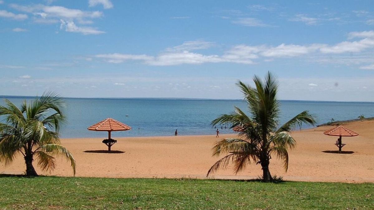 Praia de areia, coqueiros, guarda-s&oacute;is, &aacute;gua azul e grama verde sob c&eacute;u claro