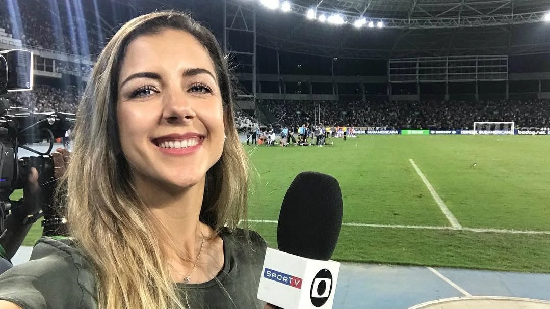 A jornalista Domitila Becker em um de seus trabalhos anteriores pelo SporTV, antes de ser contratada pela ESPN para produzir conteúdo esportivo digital. Foto: Reprodução Jornalista Domitila Becker sorrindo, segurando microfone SporTV em estádio de futebol à noite com público