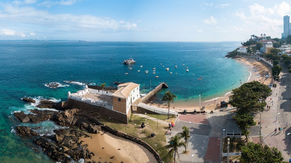 Vista a&eacute;rea de uma ba&iacute;a com forte hist&oacute;rico, praia com pessoas, mar azul com barcos e cidade ao fundo.