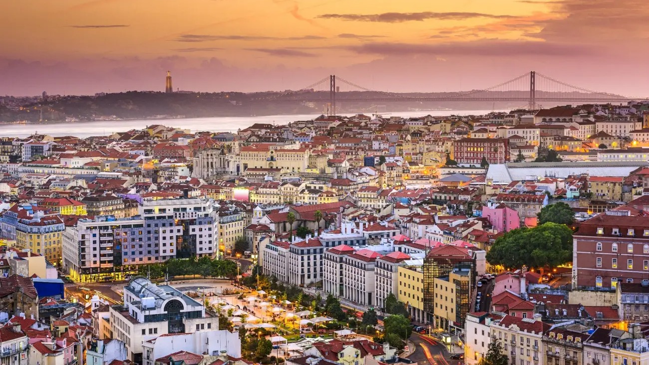 Com sua luz única e arquitetura histórica, Lisboa atrai jogadores de futebol em busca de qualidade de vida e paixão esportiva. Vista aérea de Lisboa ao entardecer, com rio, ponte, prédios e o Cristo Rei.