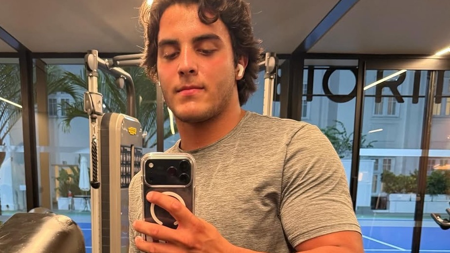 Marcelo Sangalo exibe o físico conquistado. Foto: Reprodução/Redes sociais Jovem com cabelo escuro cacheado, camiseta cinza, fones brancos, tira selfie em academia com equipamentos