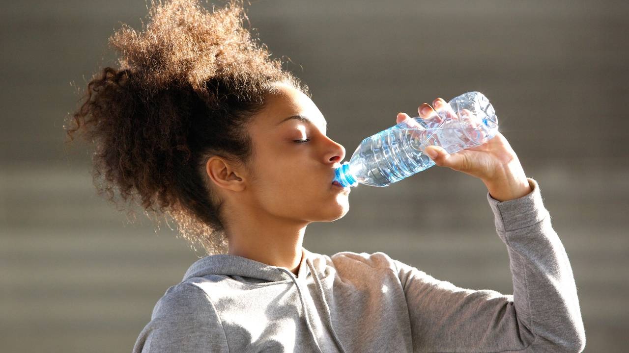 A hidratação constante é um hábito simples e eficaz para fortalecer a imunidade e proteger as vias aéreas contra doenças respiratórias no inverno. Jovem mulher com cabelo encaracolado e olhos fechados bebendo água de uma garrafa plástica.
