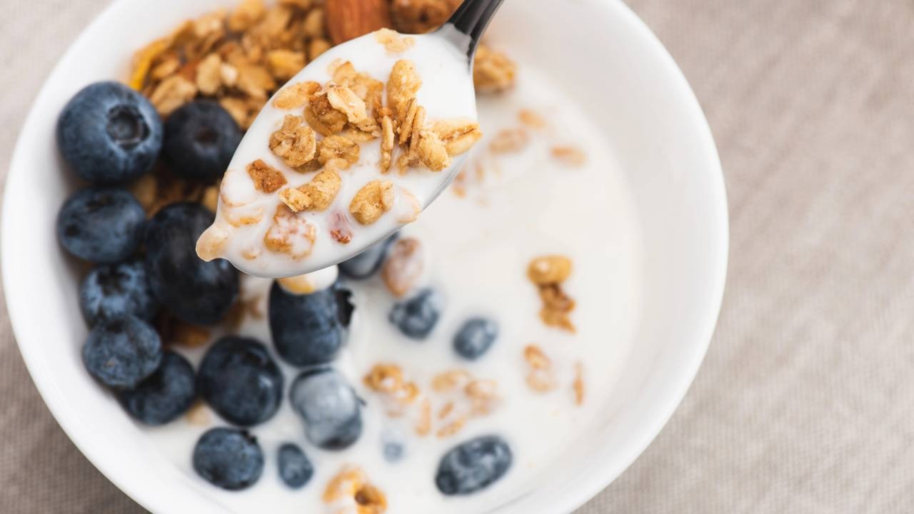 O café da manhã com iogurte e granola é uma opção prática para iniciar o dia com energia e nutrição. Colher com iogurte e granola sobre tigela de iogurte, mirtilos e mais granola