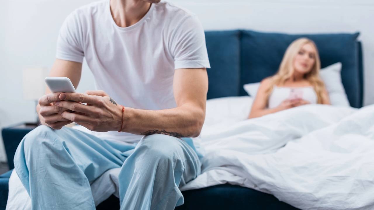 O celular no quarto: um sinal de alerta para a falta de honestidade nos relacionamentos, como aponta a matéria. Homem sentado na cama foca no celular, mulher deitada no fundo também usa o telefone.