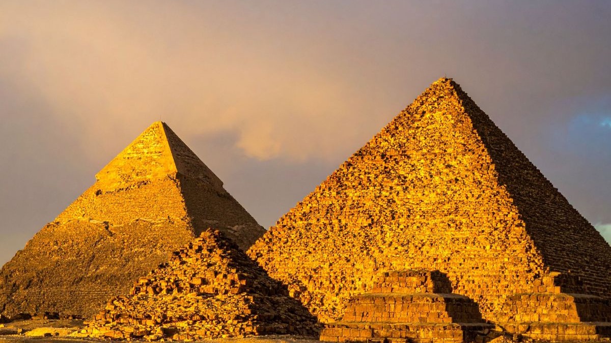Egito: com a valorização do real, as icônicas pirâmides se tornam um destino historicamente rico e financeiramente viável para brasileiros. Duas pirâmides e ruínas de pedra sob céu dourado no entardecer.