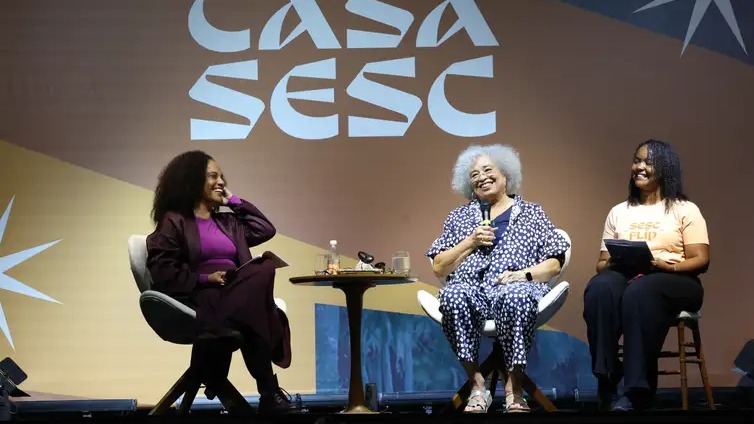Angela Davis e Flávia Oliveira em painel sobre gentrificação e cultura na 24ª Flip em Paraty. Três mulheres sorrindo em palco com "CASA SESC": uma segura microfone, outra tablet