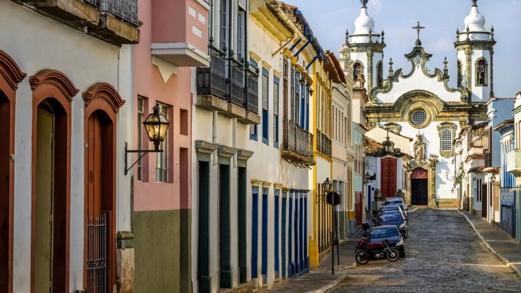 Rua de paralelepípedos ladeada por casarões coloniais coloridos e igreja barroca ao fundo.