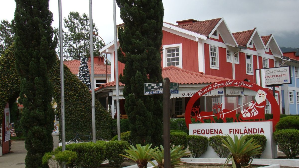 O centro comercial 'Pequena Finlândia' em Penedo, com sua Casa do Papai Noel, reflete a colonização finlandesa na serra fluminense. Entrada da Pequena Finlândia em Penedo com edifícios vermelhos, placa do Papai Noel e árvores.