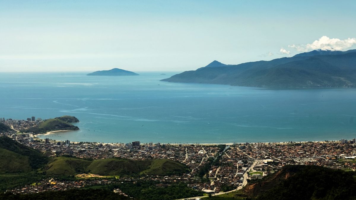 Vista aérea de uma cidade costeira com praia extensa, mar azul e ilhas ao fundo.