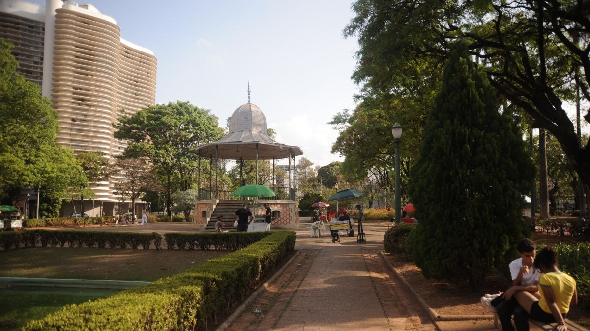 Pessoas relaxam em um parque urbano com gazebo, &aacute;rvores e um pr&eacute;dio moderno ao fundo.