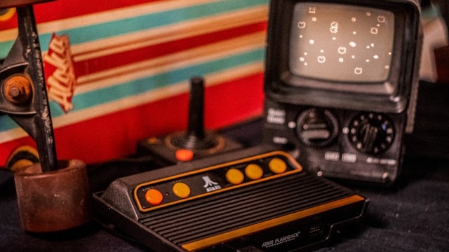 Console Atari, dois joysticks, e TV antiga com jogo pixelado, ao lado de um skate.