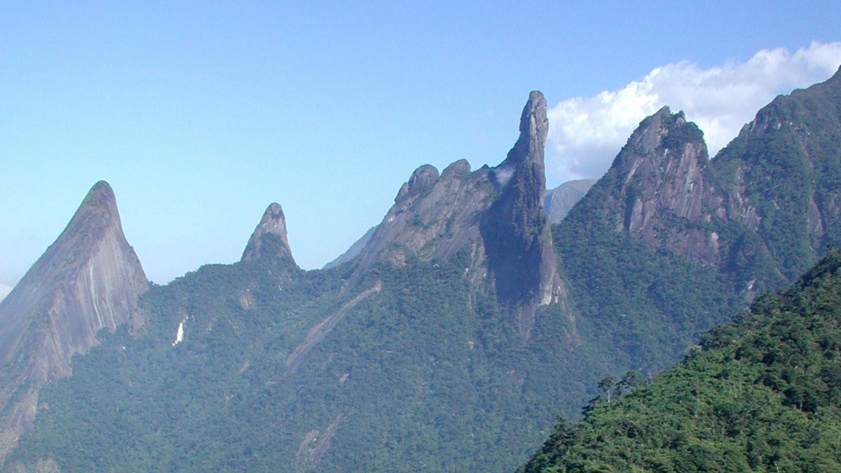 O imponente pico Dedo de Deus, na Serra dos Órgãos, convida à aventura em Teresópolis, um dos destinos da serra fluminense. Picos rochosos da Serra dos Órgãos, incluindo o Dedo de Deus, cobertos por densa floresta.